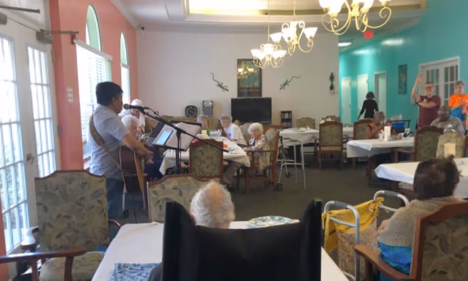 A group of elderly residents seated at tables in a dining room with white tablecloths, listening to a man playing guitar and singing into a microphone. The room has large windows with arched tops on the left, chandeliers hanging from the ceiling, and teal and pink walls. Some residents use walkers and wheelchairs.
