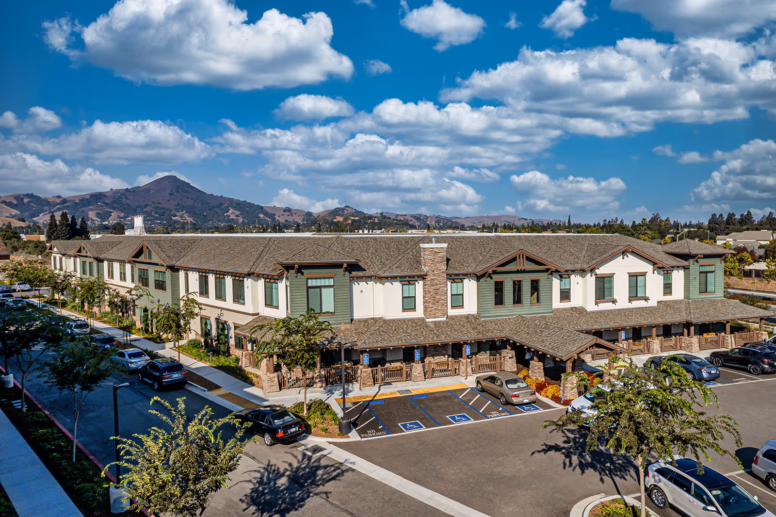 Exterior view of a two-story senior living facility building with a large parking lot in front, several cars parked, and a mountainous landscape in the background under a partly cloudy blue sky.