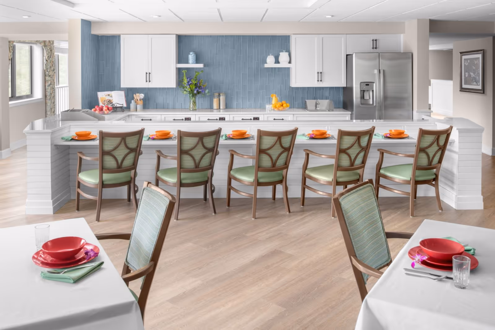 A bright and modern kitchen and dining area with a long white counter featuring six wooden chairs with green cushions. The kitchen has white cabinets, a blue tiled backsplash, a stainless steel refrigerator, and various decorative items including flowers and fruit. In the foreground, there are two dining tables set with red bowls, plates, green napkins, and glasses.