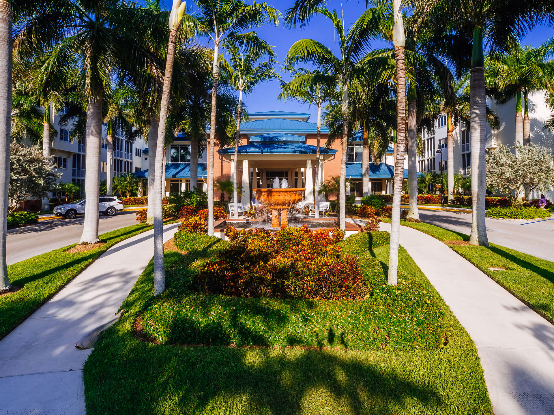 Front exterior view of The Arbor at Shell Point facility with a landscaped entrance featuring palm trees, a central fountain, and a paved walkway leading to the building entrance under a blue roof.