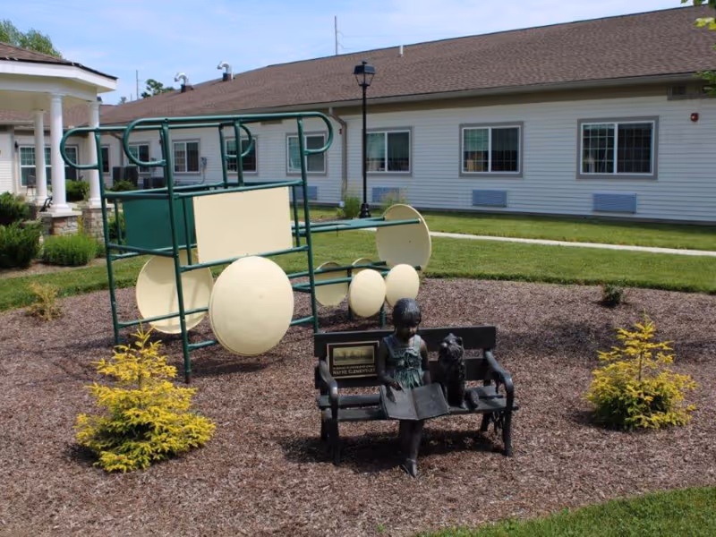 Outdoor garden area at Avenue at Wooster Care and Rehabilitation Center featuring a playground structure with circular yellow panels and a bronze statue of a girl reading a book on a bench with a dog beside her, surrounded by mulch and small shrubs, with a building in the background.