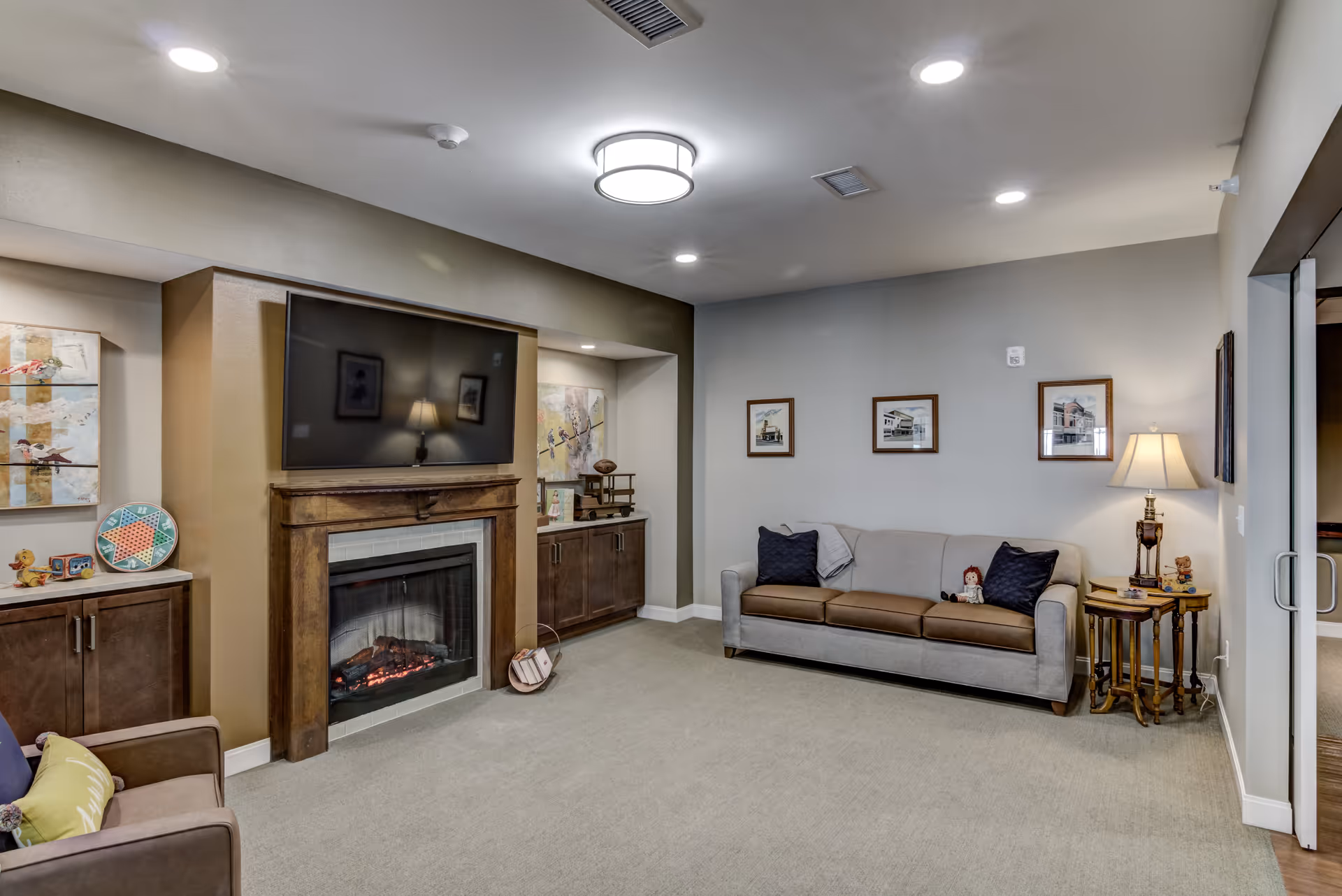A comfortable common living room with a sofa, armchair, TV mounted above a fireplace, built-in cabinets, and warm lighting.