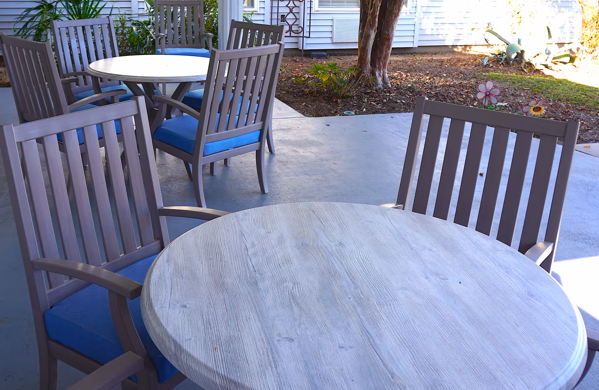 Outdoor patio area with round wooden tables and chairs with blue cushions, adjacent to a garden bed with plants and a tree, next to a building with white siding.