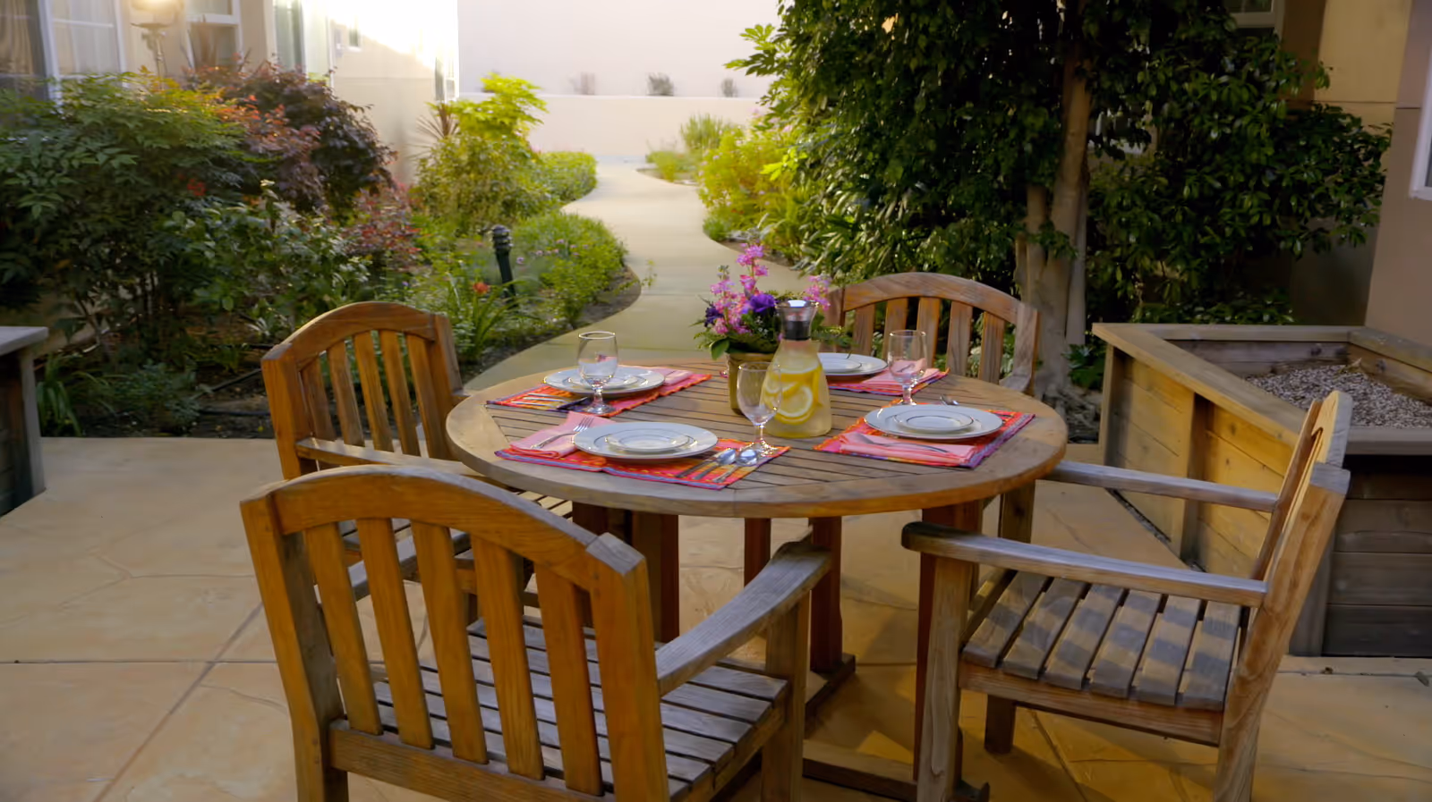 Outdoor patio area with a round wooden table set for four with plates, glasses, and a pitcher of lemon water. The table is surrounded by four wooden chairs and there are plants and greenery in the background along a curved pathway.