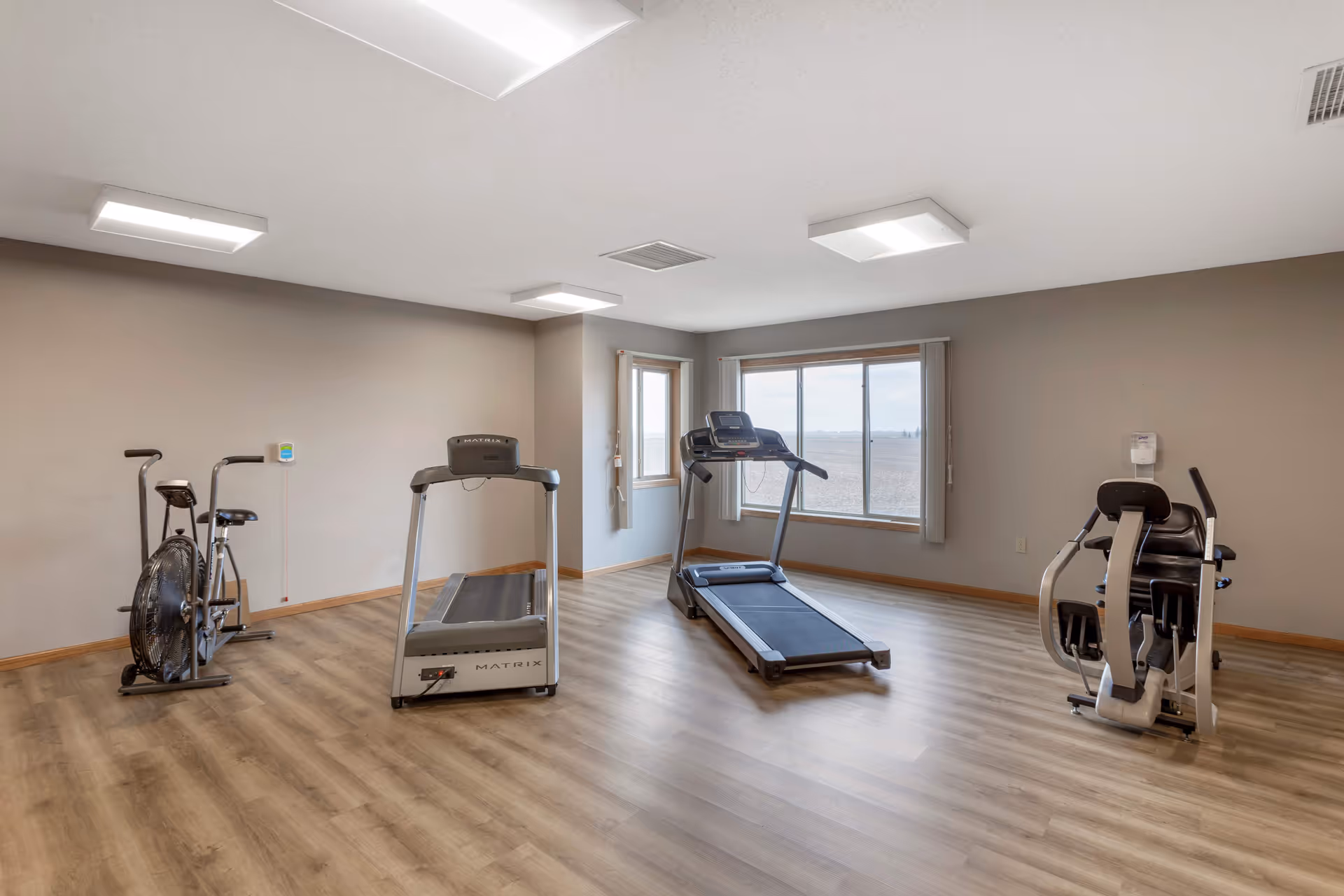Empty fitness room with a treadmill, stationary bike, and strength machine, wood-look flooring, and large windows.