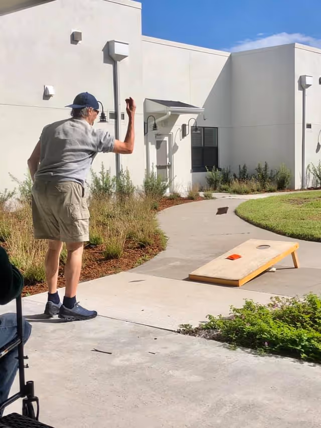 An elderly man wearing a cap, gray shirt, and khaki shorts is playing cornhole outdoors on a paved pathway next to a white building with windows and outdoor lights. There is greenery and landscaping around the pathway, and a wheelchair is partially visible in the foreground.