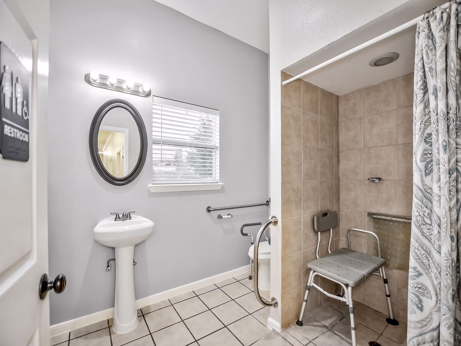 A clean, accessible restroom with a white pedestal sink, oval mirror, and a window with blinds. The restroom features a tiled shower area with a shower chair and grab bars, a toilet with grab bars, and a patterned shower curtain.
