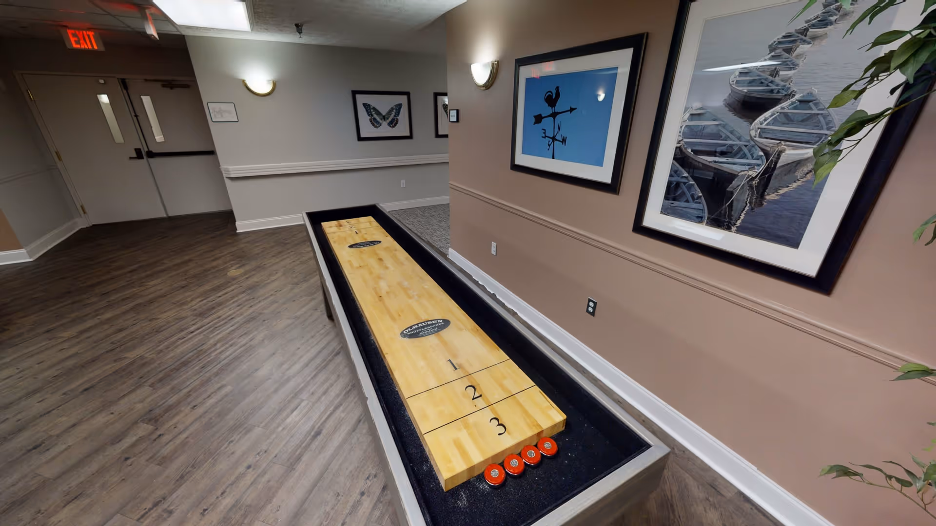 Indoor recreational area with a shuffleboard table on a wood floor. The walls are decorated with framed pictures, including a butterfly, a weather vane, and boats docked on water. There is an exit door in the background.
