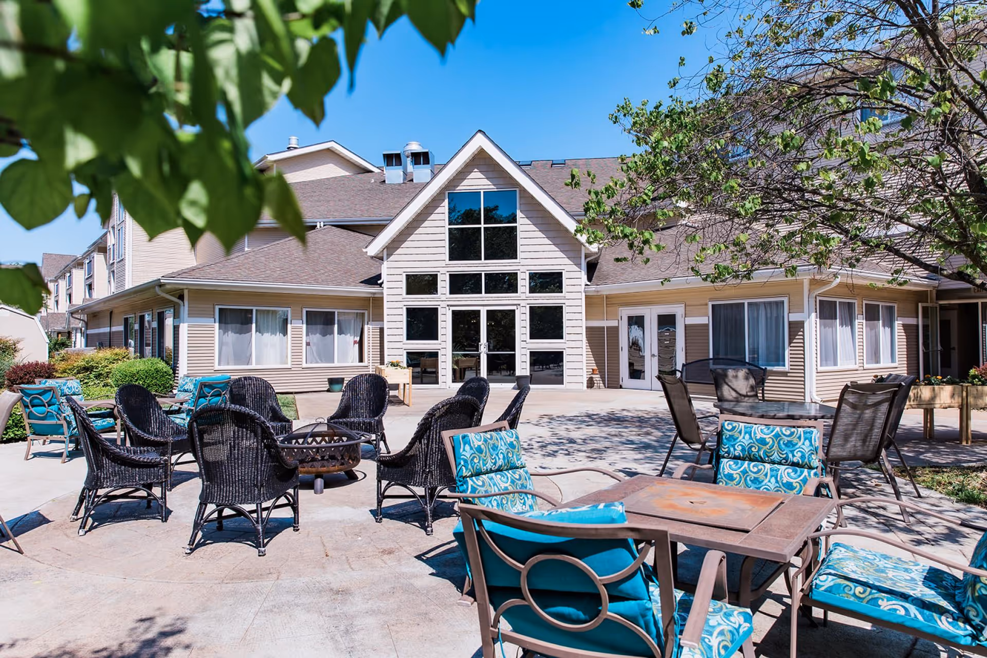 Outdoor patio area at College View Manor with multiple seating arrangements including wicker chairs around a fire pit and cushioned chairs around tables, surrounded by trees and greenery under a clear blue sky.