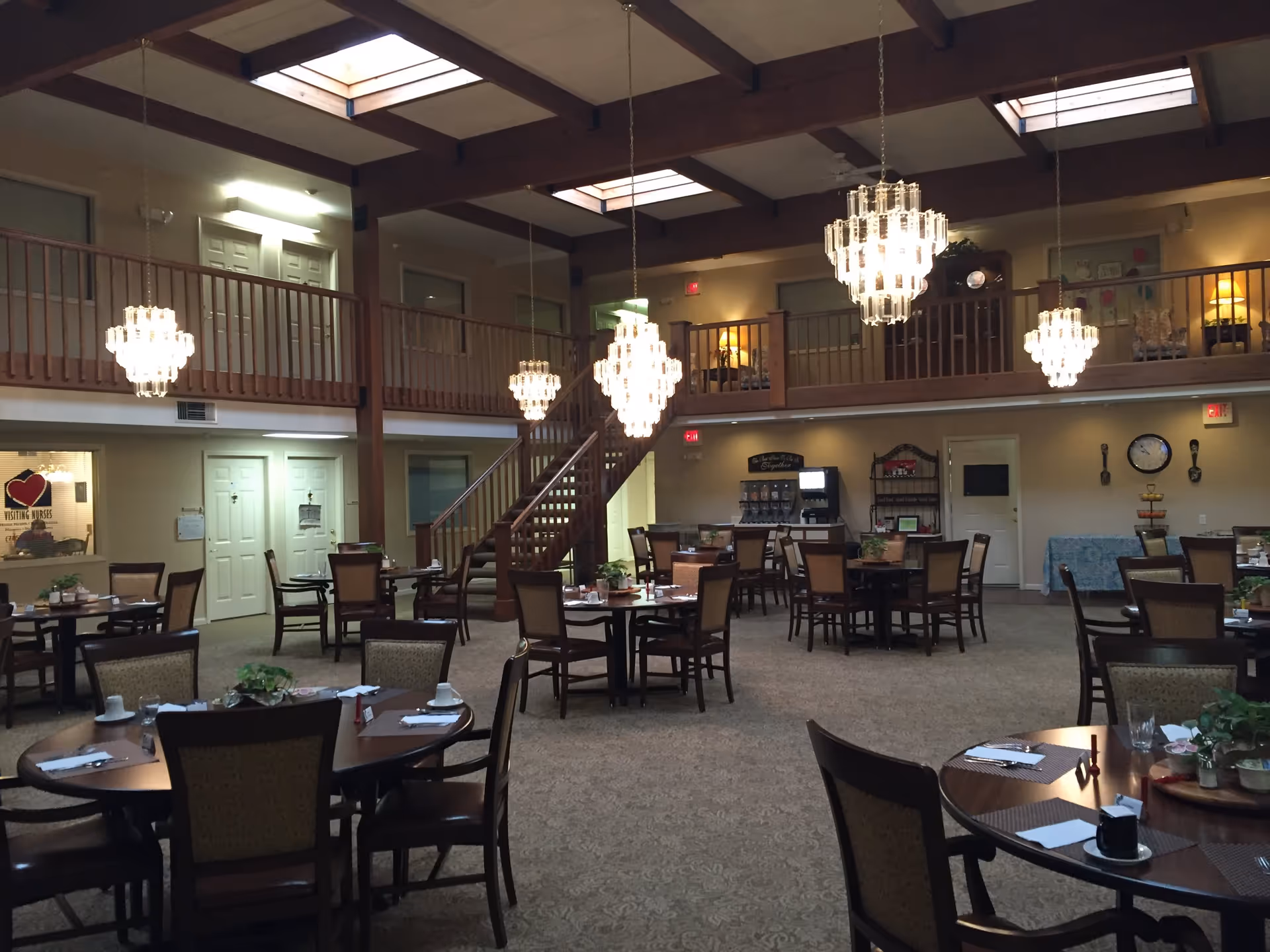 A spacious dining room in a retirement community with multiple round tables and chairs arranged neatly. The room features a high ceiling with wooden beams and skylights, elegant chandeliers hanging from the ceiling, and a staircase leading to an upper balcony area. The walls have doors and windows, and there is a beverage station against one wall.
