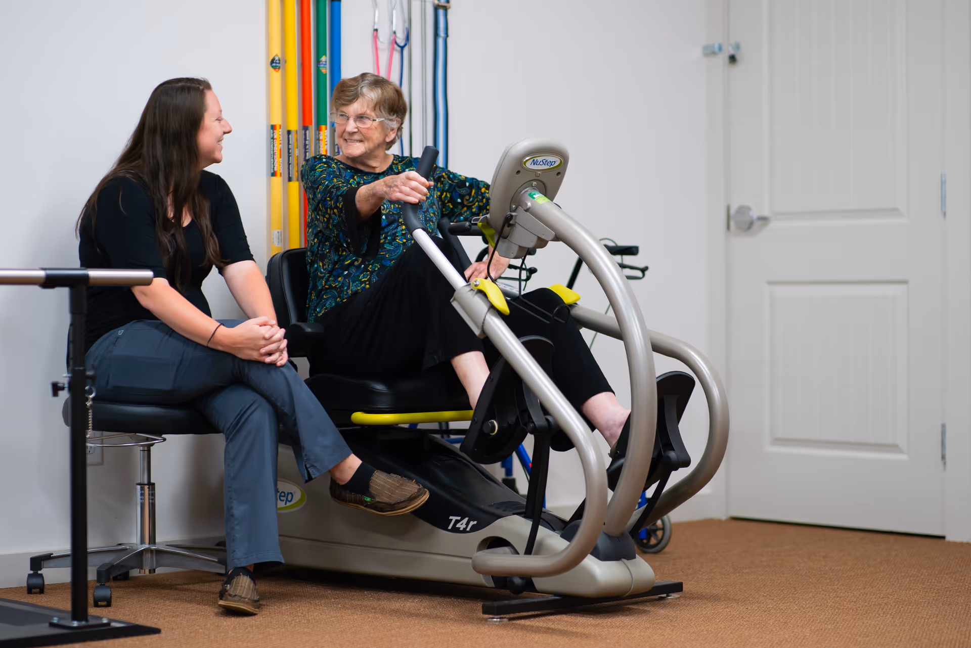 An elderly woman using a NuStep exercise machine in a room, smiling and interacting with a younger woman sitting beside her on a chair. Various colorful exercise poles are visible in the background, along with a closed white door.