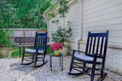 Outdoor patio area with two black rocking chairs and a small black table between them holding a pot of pink flowers. The patio is paved with stone tiles and is adjacent to a light-colored building wall. There is a wooden fence and green trees in the background.