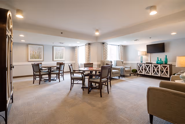 Bright senior living common room with round tables and chairs, armchairs, a TV on a console, and windows with patterned curtains.