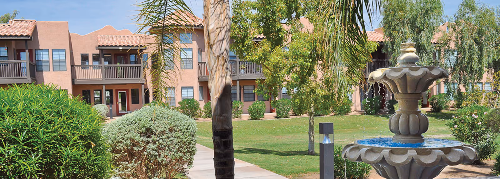 Outdoor view of a senior living facility with a two-story building featuring balconies and red-tiled roofs. The foreground shows a landscaped garden with green bushes, trees, a walking path, and a decorative water fountain.