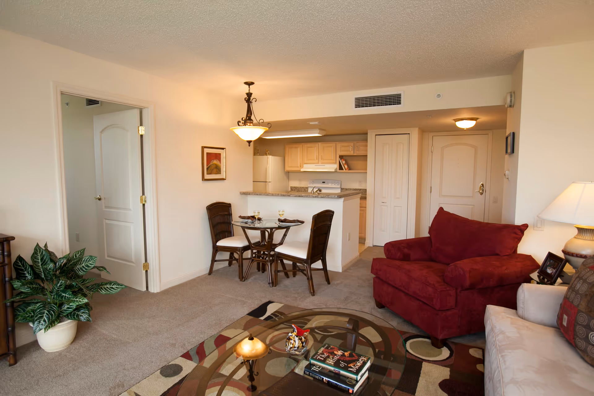 Interior view of a senior living facility apartment at Mangrove Bay showing a cozy living room with a red armchair, beige sofa, glass coffee table with books, a small dining area with a round glass table and two chairs, and a kitchen with light wood cabinets and white appliances in the background.