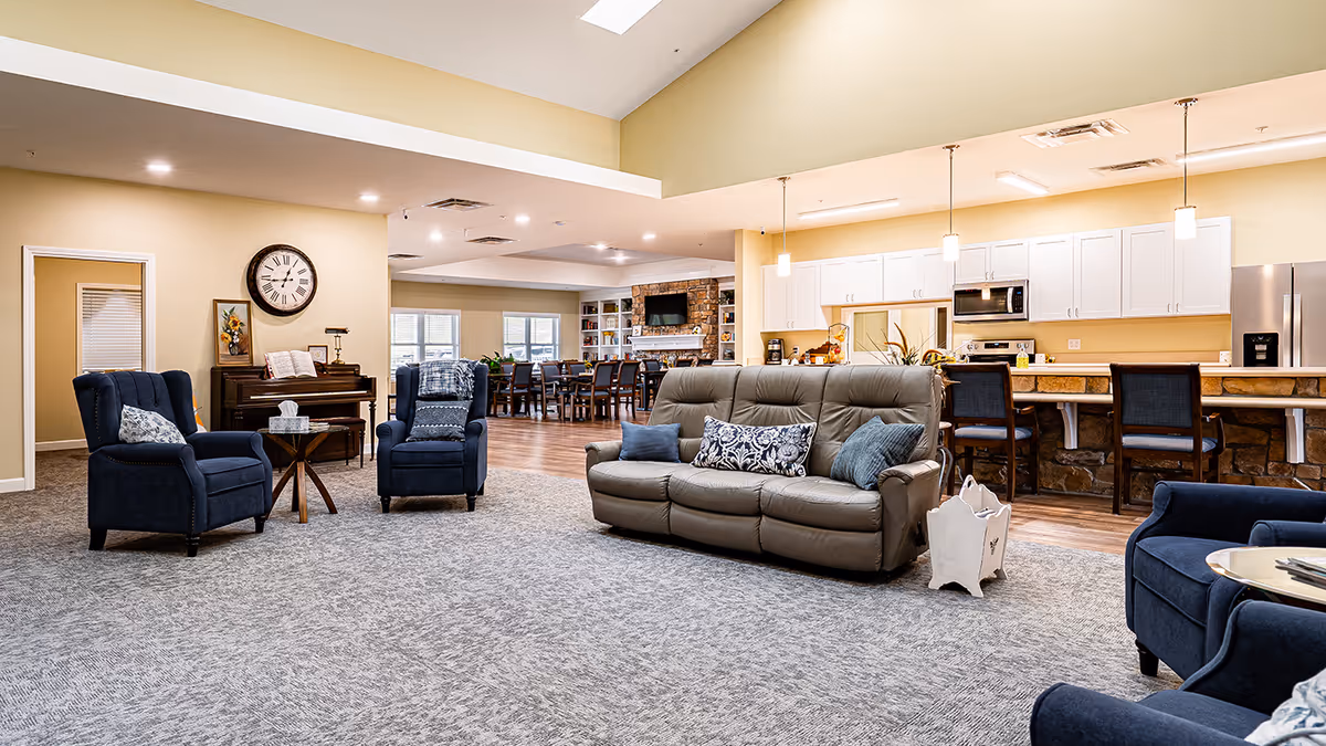 A spacious and well-lit common living area in a senior living facility featuring a gray leather sofa with decorative pillows, two blue armchairs, a piano with a clock above it, and a kitchen area with white cabinets and bar stools in the background.