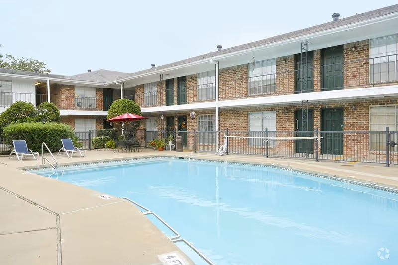 Outdoor swimming pool area at Emerson Pines with clear blue water, surrounded by a concrete deck with lounge chairs and a table with a red umbrella. The pool is fenced and adjacent to a two-story brick building with green doors and windows.