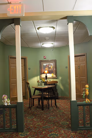 Carpeted interior common area with a round table and chairs, a table lamp under a framed painting, flanked by doors and decorative columns.
