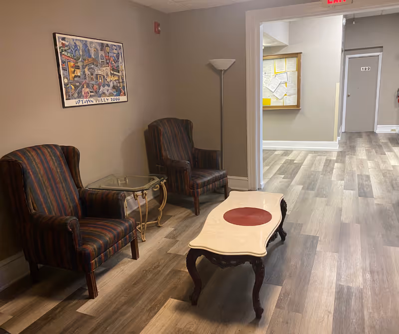 A small sitting area with two striped upholstered armchairs, a glass side table, and a decorative coffee table with a red circular centerpiece. The room has wood-patterned flooring, beige walls, a floor lamp, and a colorful framed artwork on the wall. In the background, there is a hallway with a bulletin board and a door labeled '100'.