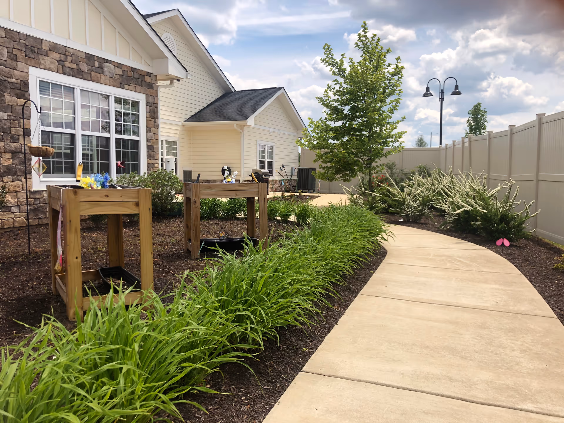 Curved concrete pathway beside a building with stone and light yellow siding exterior walls, bordered by green plants and shrubs, with two wooden raised garden beds and a tree near a white fence under a partly cloudy sky.
