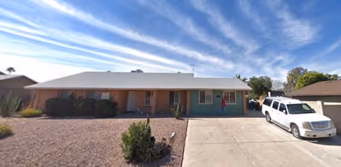 Single-story residential building with a brick and green exterior, a driveway with a white SUV parked, and a clear blue sky with wispy clouds above.
