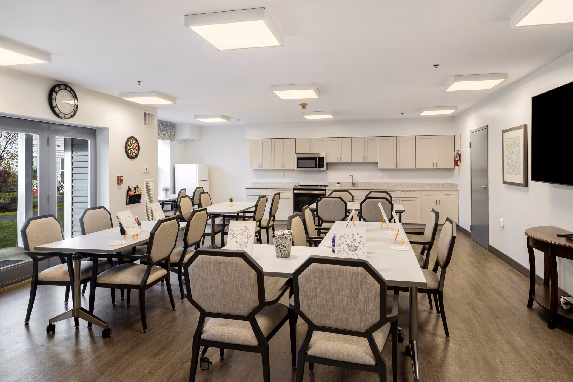 A bright and spacious communal room with several tables and cushioned chairs arranged for group activities. The tables have coloring sheets and art supplies on them. The room features a kitchenette with white cabinets, a microwave, and a refrigerator. There is a large wall-mounted TV, a clock, a dartboard, and glass doors leading outside to a garden area.