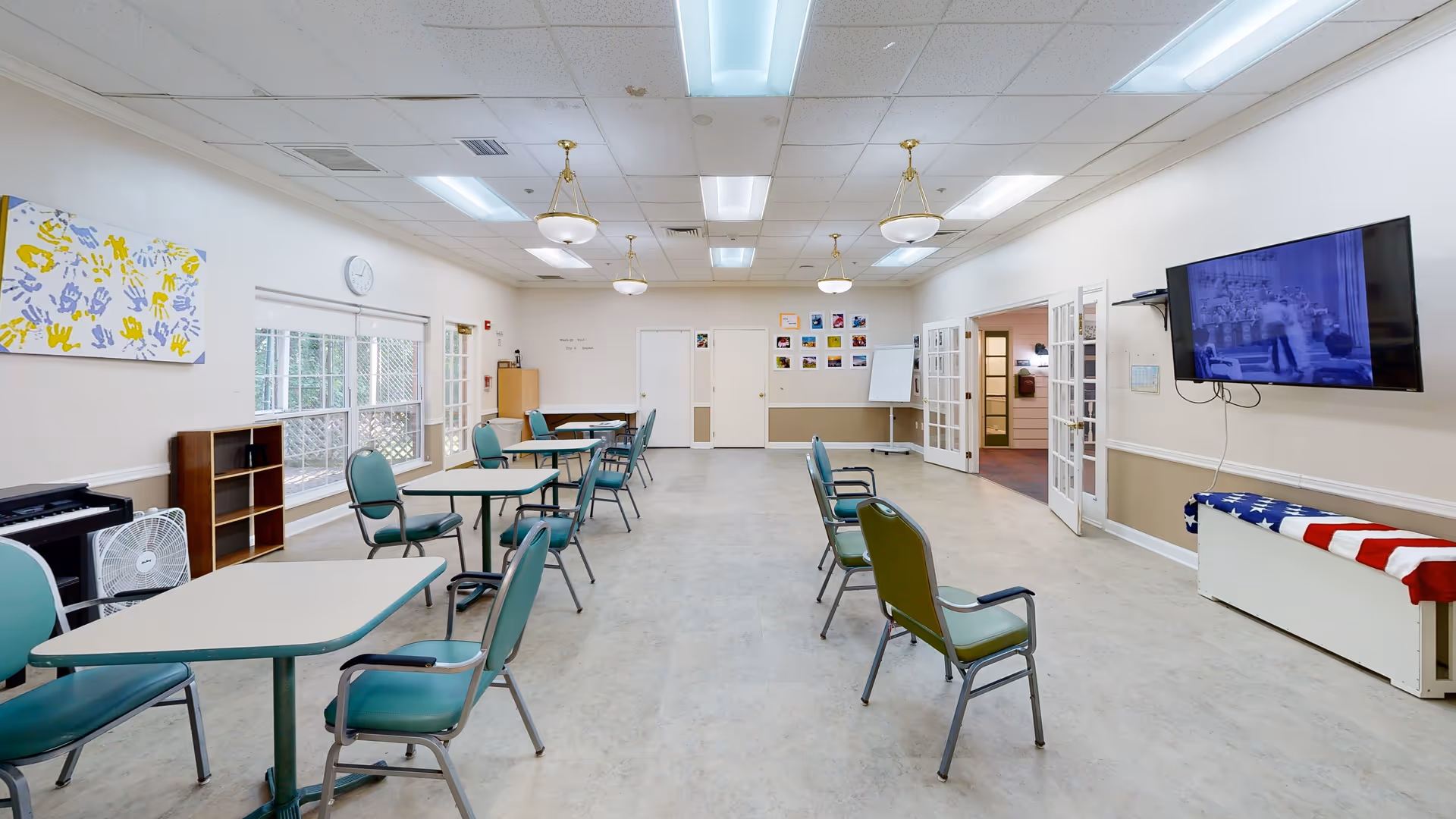A spacious room with several tables and green chairs arranged with social distancing. The room has white walls with beige wainscoting, a large flat-screen TV mounted on the right wall, and a bench covered with an American flag blanket beneath it. There are windows on the left side, a clock above the windows, and colorful handprint artwork on the left wall. The ceiling has fluorescent lights and hanging light fixtures. Double glass doors are open on the right side, leading to another room.