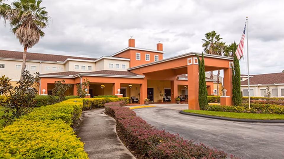 Front entrance of a coral-orange assisted living building with a covered porte-cochère, landscaped driveway, palm trees, and an American flag.