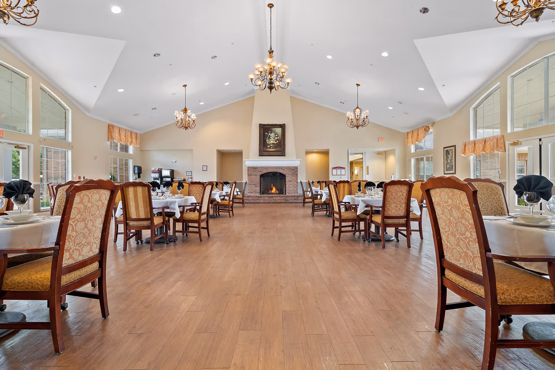 Spacious dining room with multiple round tables covered with white tablecloths, each set with plates, glasses, and black napkins. The room features wooden chairs with patterned upholstery, a wooden floor, large windows with orange valances, chandeliers hanging from a high vaulted ceiling, and a brick fireplace with a framed painting above it at the far end.