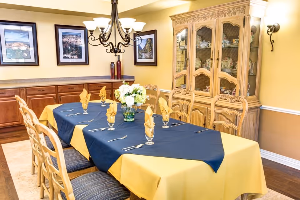 A dining room with a rectangular table covered with a yellow tablecloth and a navy blue overlay. The table is set with six glasses each holding a folded yellow napkin, and silverware is placed beside each setting. A vase with white flowers is centered on the table. Wooden chairs with cushioned seats surround the table. The room has yellow walls with framed landscape photographs, a wooden cabinet displaying china, and a chandelier hanging above the table.
