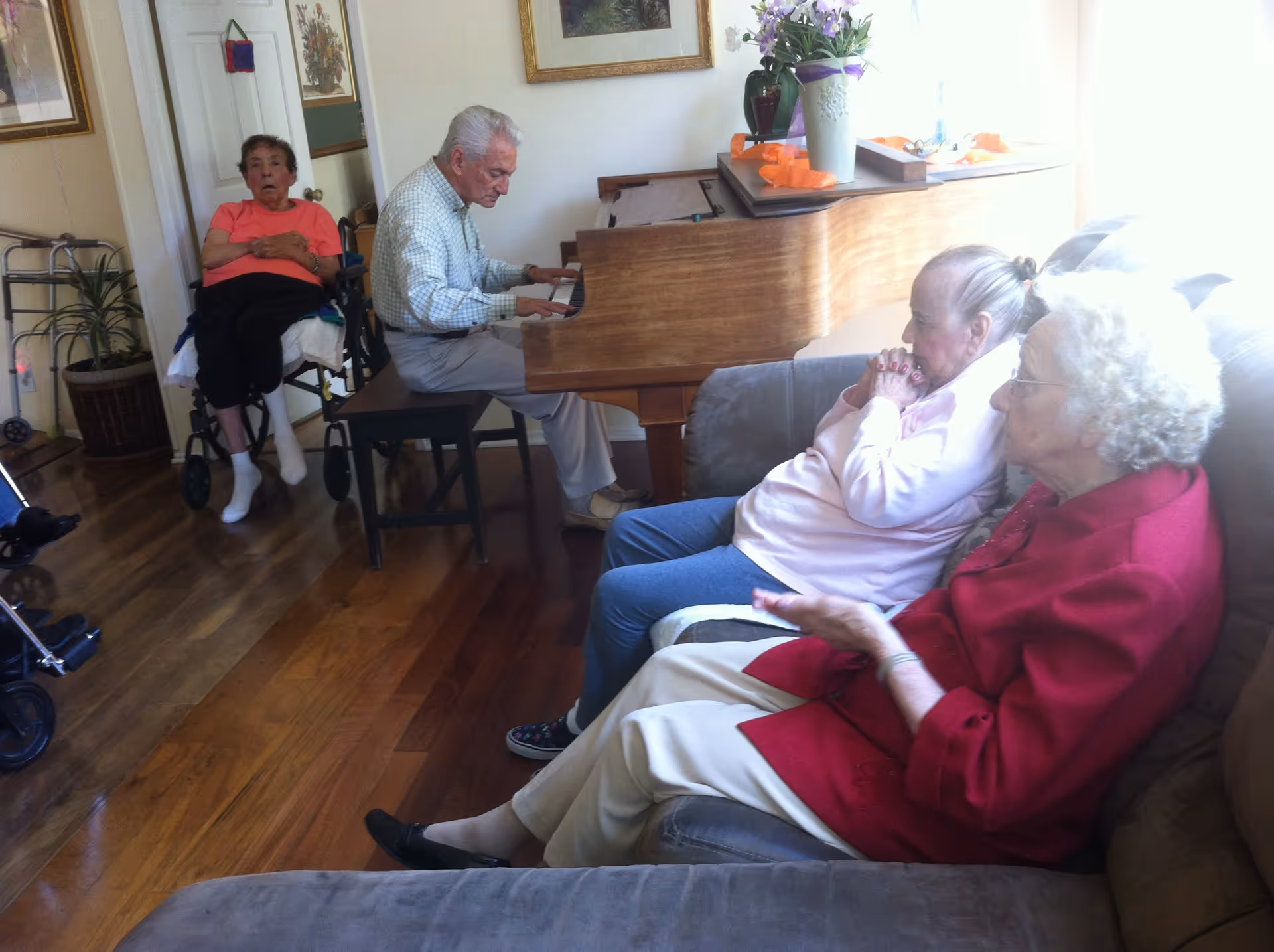 An elderly man playing a wooden piano in a living room while three elderly women sit nearby, two on a couch and one in a wheelchair, listening. The room has wooden floors, framed artwork on the walls, and a vase with flowers on the piano.