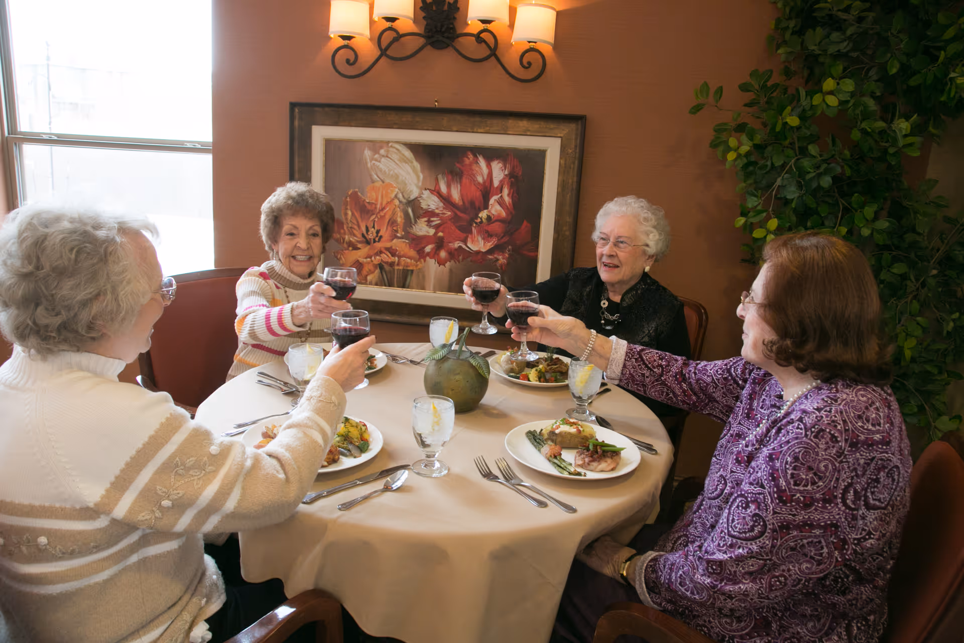 Four elderly women sitting around a round dining table in a warmly decorated room, raising glasses of red wine in a toast. The table is set with plates of food, glasses of water with lemon, and silverware. A framed floral painting and a wall sconce are visible on the wall behind them, and a large green plant is in the corner.