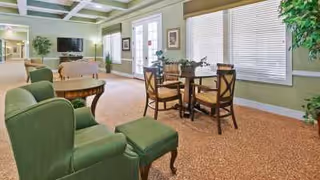 A spacious and well-lit common area in an assisted living facility featuring a green upholstered armchair with matching ottoman, a wooden table with four chairs, large windows with blinds, a flat-screen TV mounted on the wall, and decorative plants.