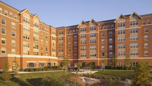 Exterior view of a multi-story brick senior living facility building with multiple windows, surrounded by landscaped greenery and small trees under a clear sky.