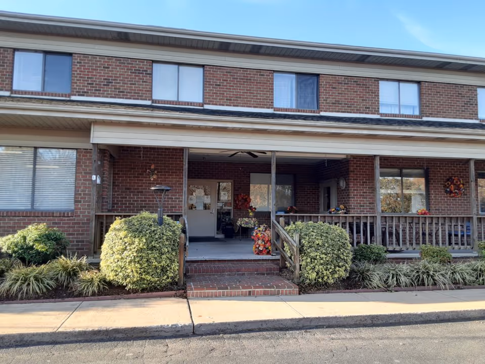 Front entrance of a two-story brick building with a covered porch, steps, shrubs, and seasonal decorations.