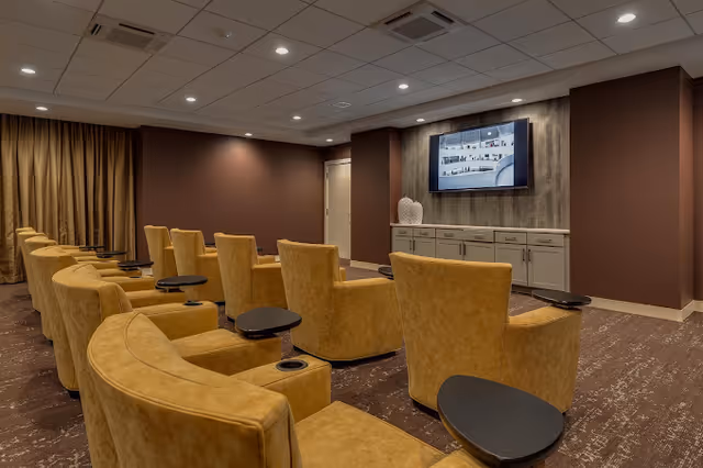 A cozy media room with multiple mustard yellow armchairs arranged in rows facing a wall-mounted flat screen TV. Each chair has a small black side table attached. The room has brown walls, a patterned carpet, recessed ceiling lights, and a cabinet beneath the TV with decorative items on top.