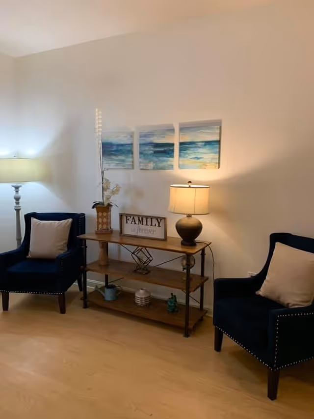 A cozy living room area with two dark blue armchairs, each with a light-colored pillow. Between the chairs is a wooden console table with three shelves, decorated with a table lamp, a vase with flowers, a decorative sign that reads 'FAMILY is forever', and various small decorative items. Above the table are three wall art pieces depicting an ocean scene. The room has light-colored walls and wooden flooring.