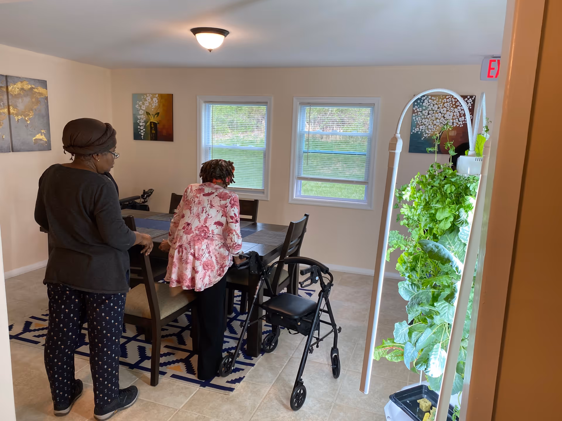 Two people stand at a dining table in a bright common room with a walker, chairs, and a tall indoor plant tower near the window.