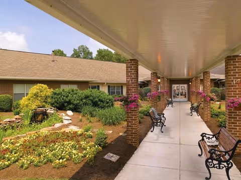 Covered outdoor walkway with benches and hanging flower baskets leading to the entrance of a single-story building surrounded by landscaped gardens.