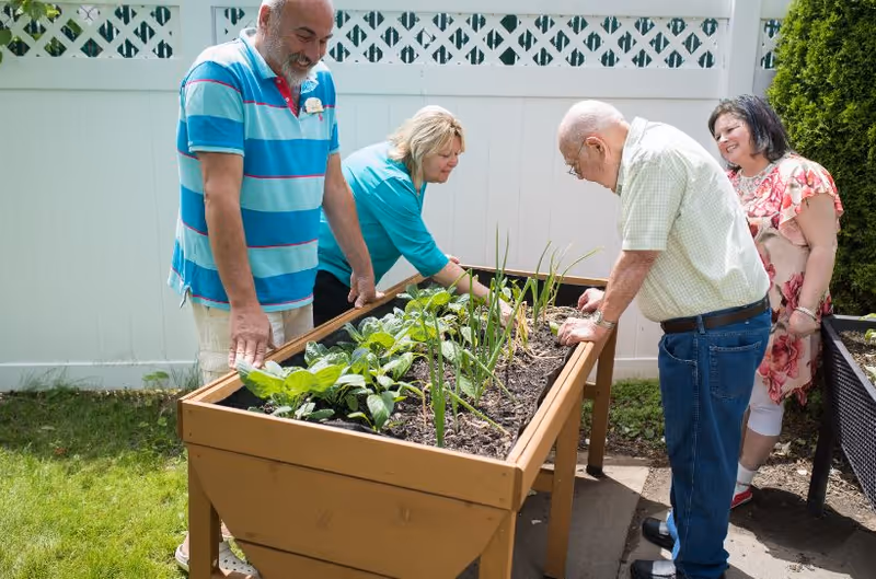 Four adults, two men and two women, tending to plants in a raised garden bed outdoors next to a white fence. They appear engaged and happy while gardening.