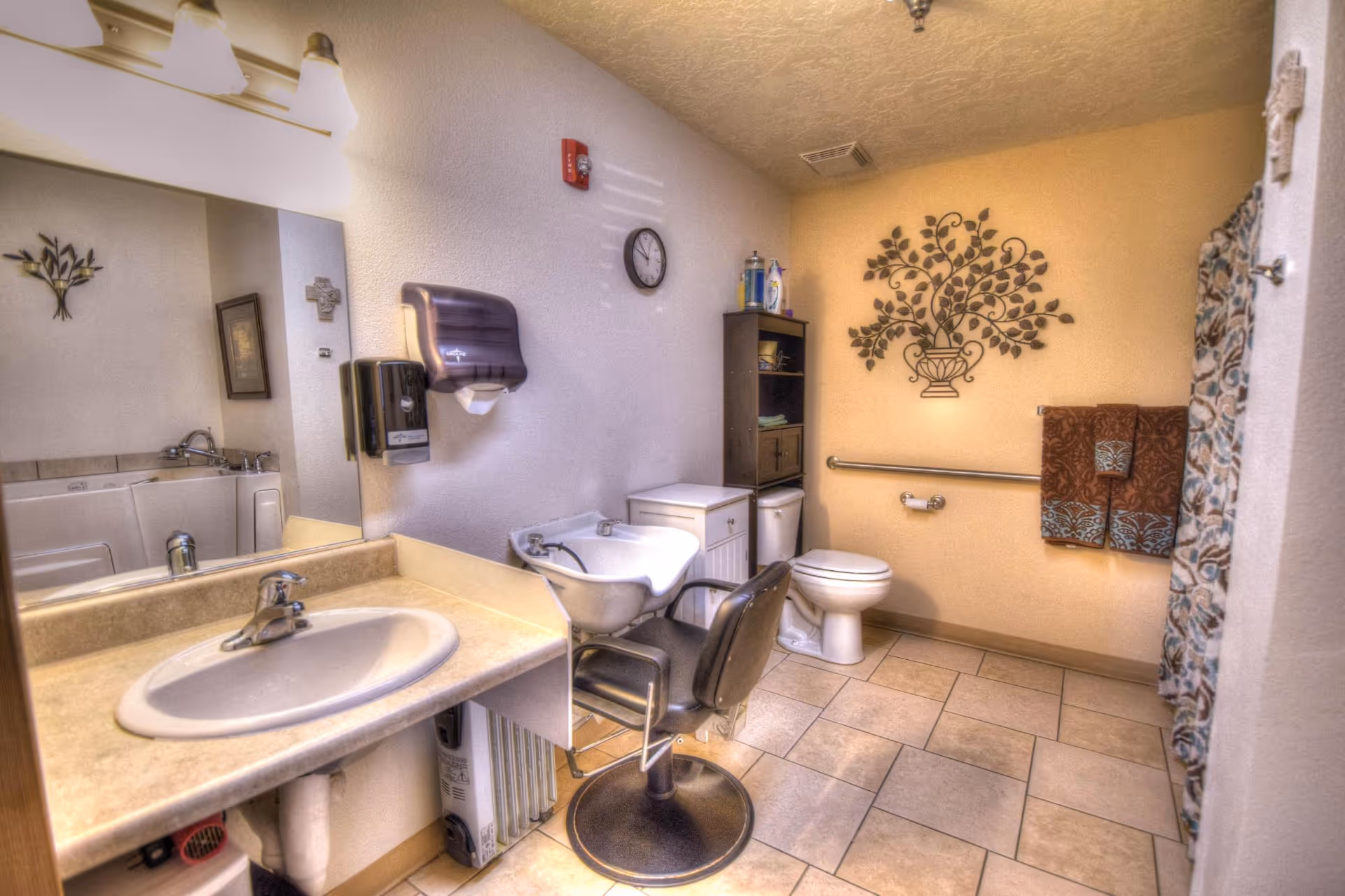 A bathroom with a sink and countertop on the left, a large mirror above the sink, a hair washing station with a chair in the center, a toilet against the far wall, and a shower with a patterned curtain on the right. The walls are decorated with a metal wall art piece of a tree and a framed picture. There are towels hanging on a bar next to the toilet, and a small shelving unit with toiletries and supplies.
