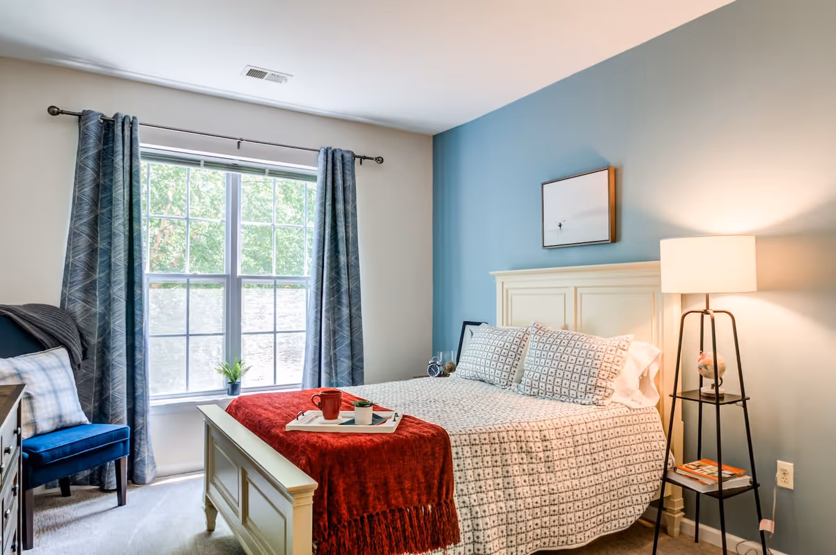 A bright bedroom with a large window covered by blue patterned curtains. The room features a white wooden bed with patterned bedding and a red throw blanket. A small tray with a red mug and a small plant sits on the bed. To the right of the bed is a tall black floor lamp with a white shade and a small globe on a lower shelf. A blue accent wall behind the bed has a framed picture hanging on it. A blue upholstered chair with a plaid pillow and a gray throw blanket is positioned near the window.