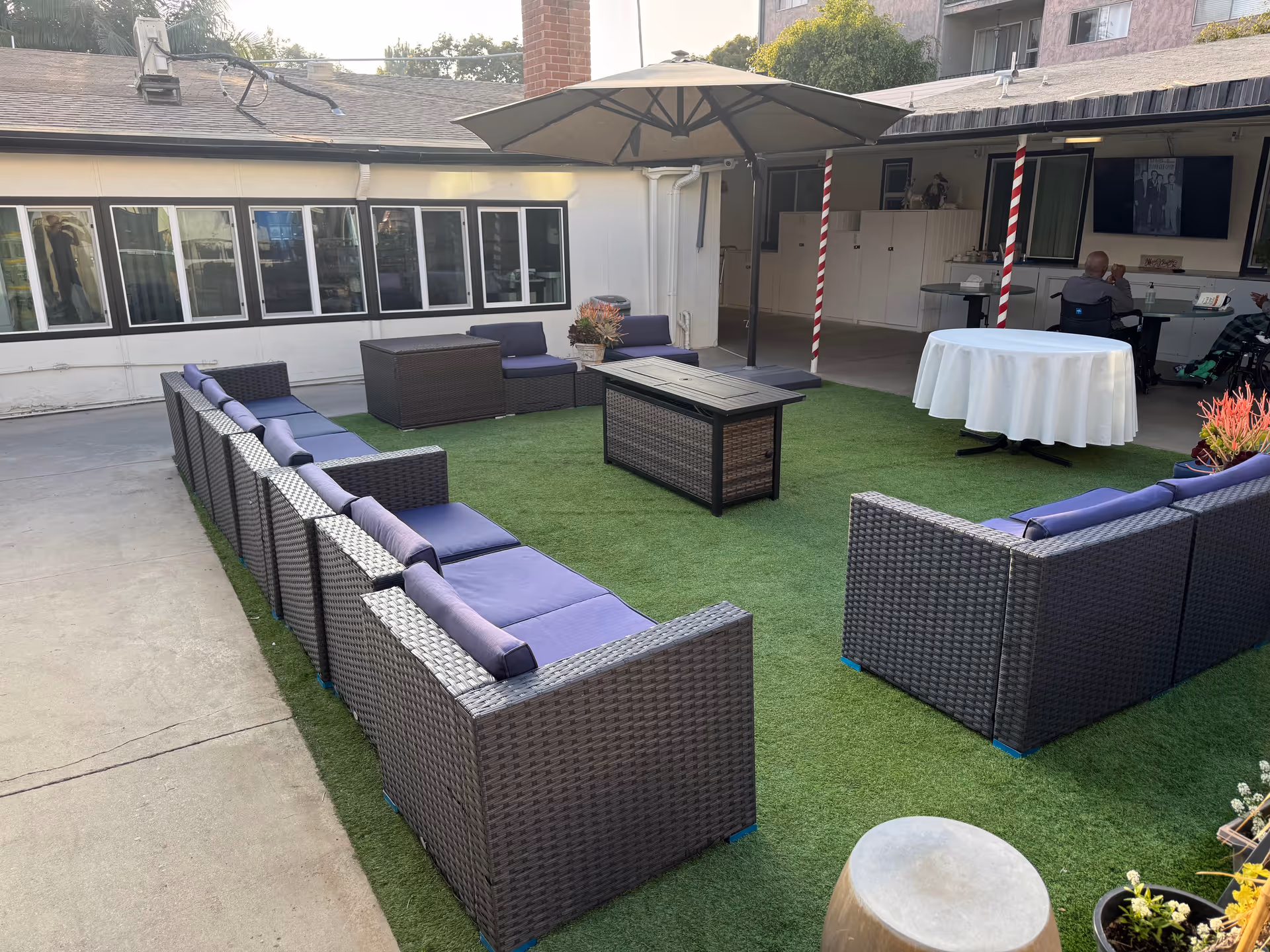 Outdoor seating area at Fireside Health Care Center with a large U-shaped arrangement of wicker chairs with purple cushions around a central rectangular table on artificial grass. There is a large umbrella providing shade, a round table covered with a white tablecloth, and two people sitting in the background near a building with multiple windows.