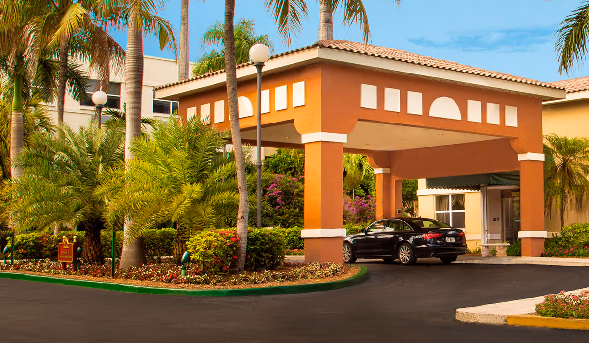 Entrance of The Palace Nursing and Rehabilitation Center featuring a covered driveway with a car parked underneath, surrounded by palm trees and well-maintained landscaping under a clear blue sky.