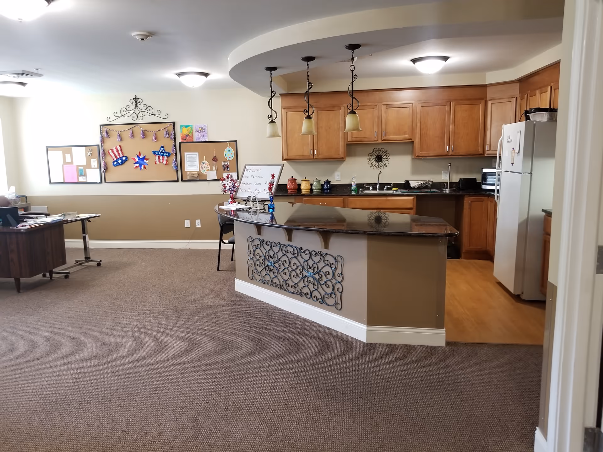 Interior view of a senior living facility kitchen and office area. The kitchen features wooden cabinets, a refrigerator, a sink, and a countertop with three hanging pendant lights. There is a decorative metal panel on the front of the counter. To the left, there is an office desk with papers and a chair. The walls have bulletin boards with various decorations and notices.