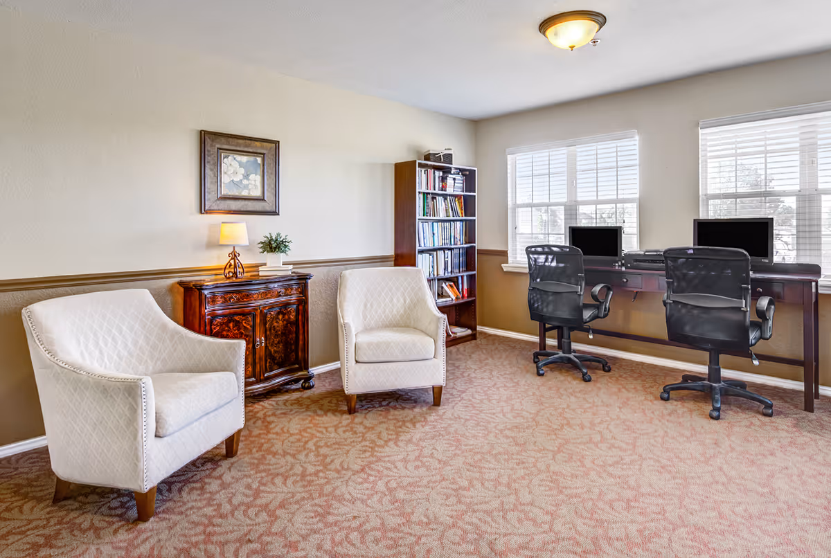 A cozy interior room with two white upholstered armchairs, a wooden cabinet with a small lamp and a framed floral picture above it, a tall bookshelf filled with books, and a long desk with two computer monitors and two black office chairs in front of windows with white blinds.