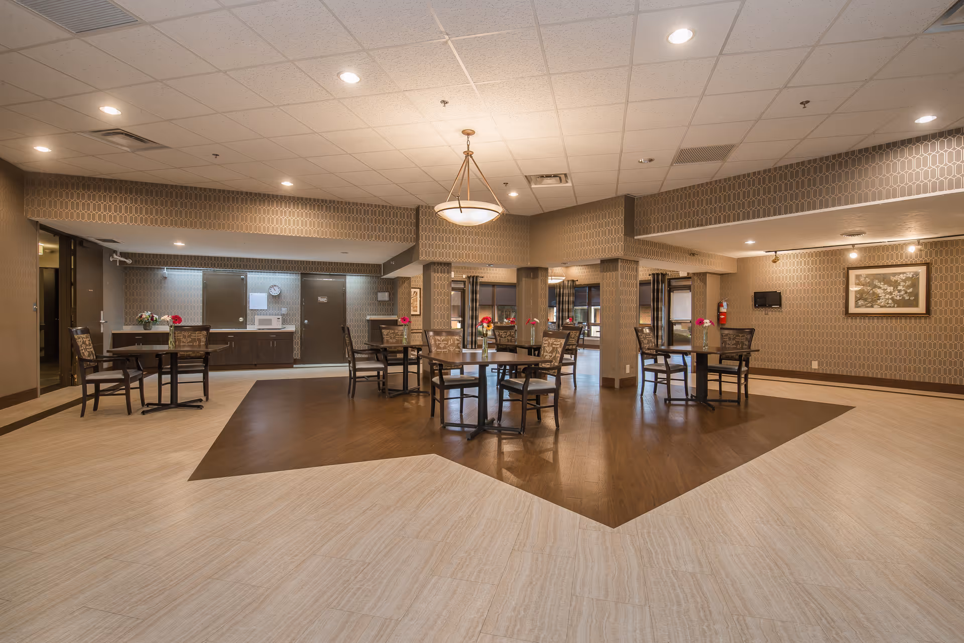 A spacious dining area in a senior living facility with several round tables and chairs arranged on a wooden floor section surrounded by a lighter tiled floor. The walls have a patterned wallpaper, and there are ceiling lights and a hanging light fixture. The background includes a kitchenette area with cabinets, a clock, and doors.