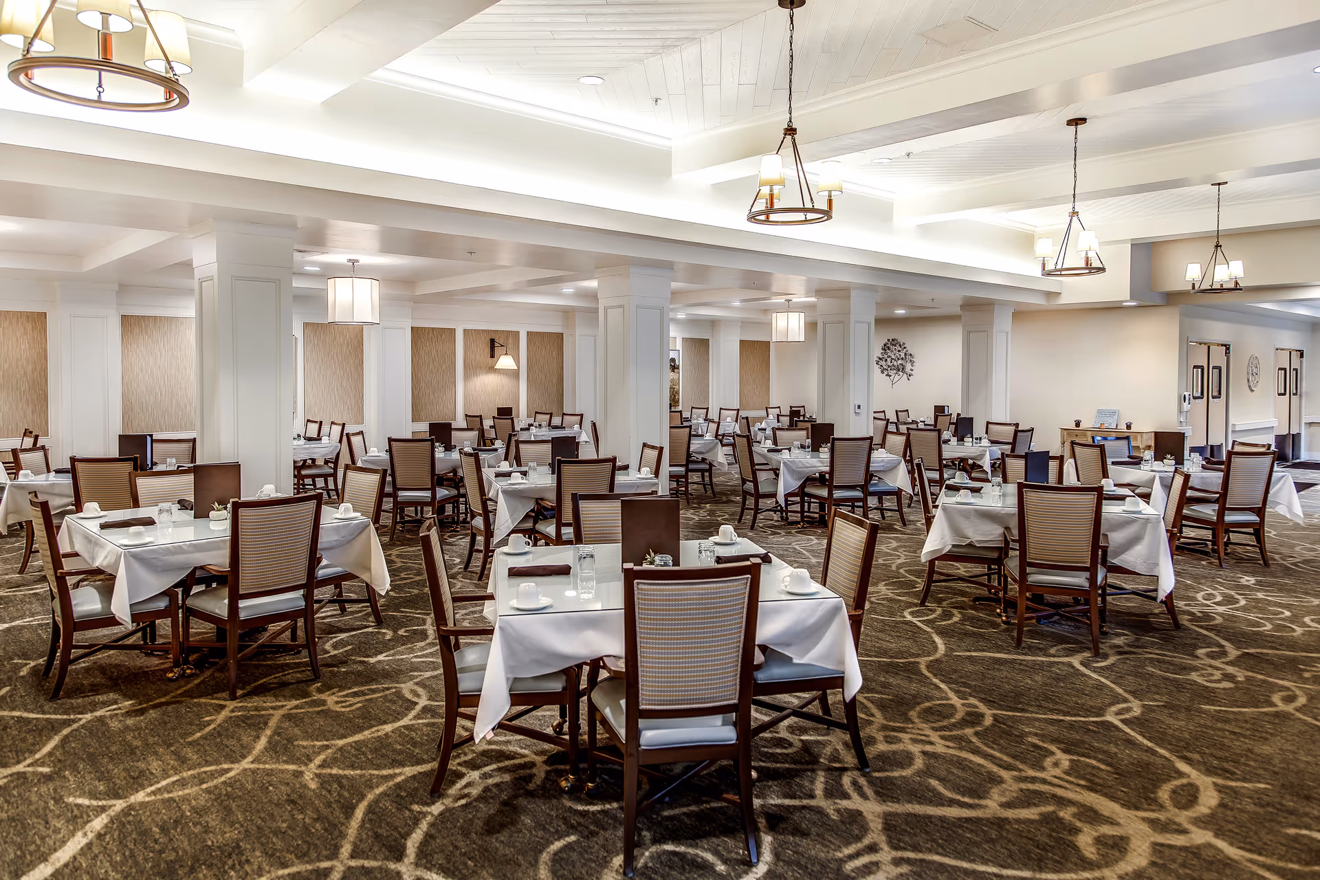 A spacious dining room with multiple tables covered in white tablecloths, each set with cups, glasses, and napkins. The room features patterned carpet, white walls with decorative panels, and several ceiling light fixtures providing warm lighting.