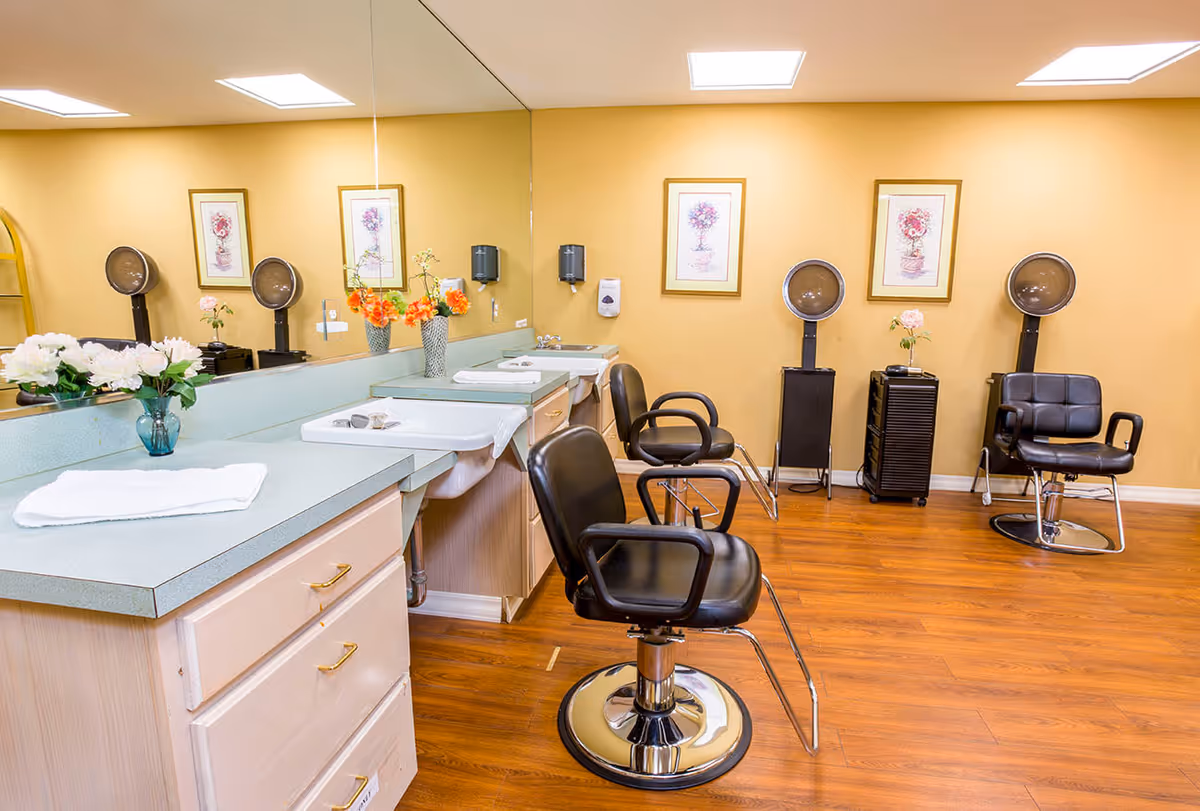 Interior view of a senior living facility's hair salon area with three black salon chairs, two hair dryers, a countertop with sinks, flowers in vases, and framed floral artwork on the yellow walls.
