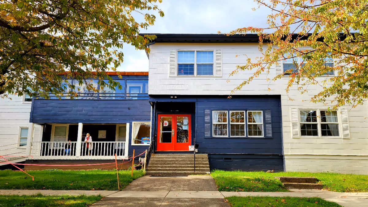 Front exterior view of a two-story building with white and dark blue siding, red double doors at the entrance, and several windows. There is a small set of stairs leading up to the entrance, a porch area with a railing, and a person standing on the porch. Trees with autumn-colored leaves frame the scene.